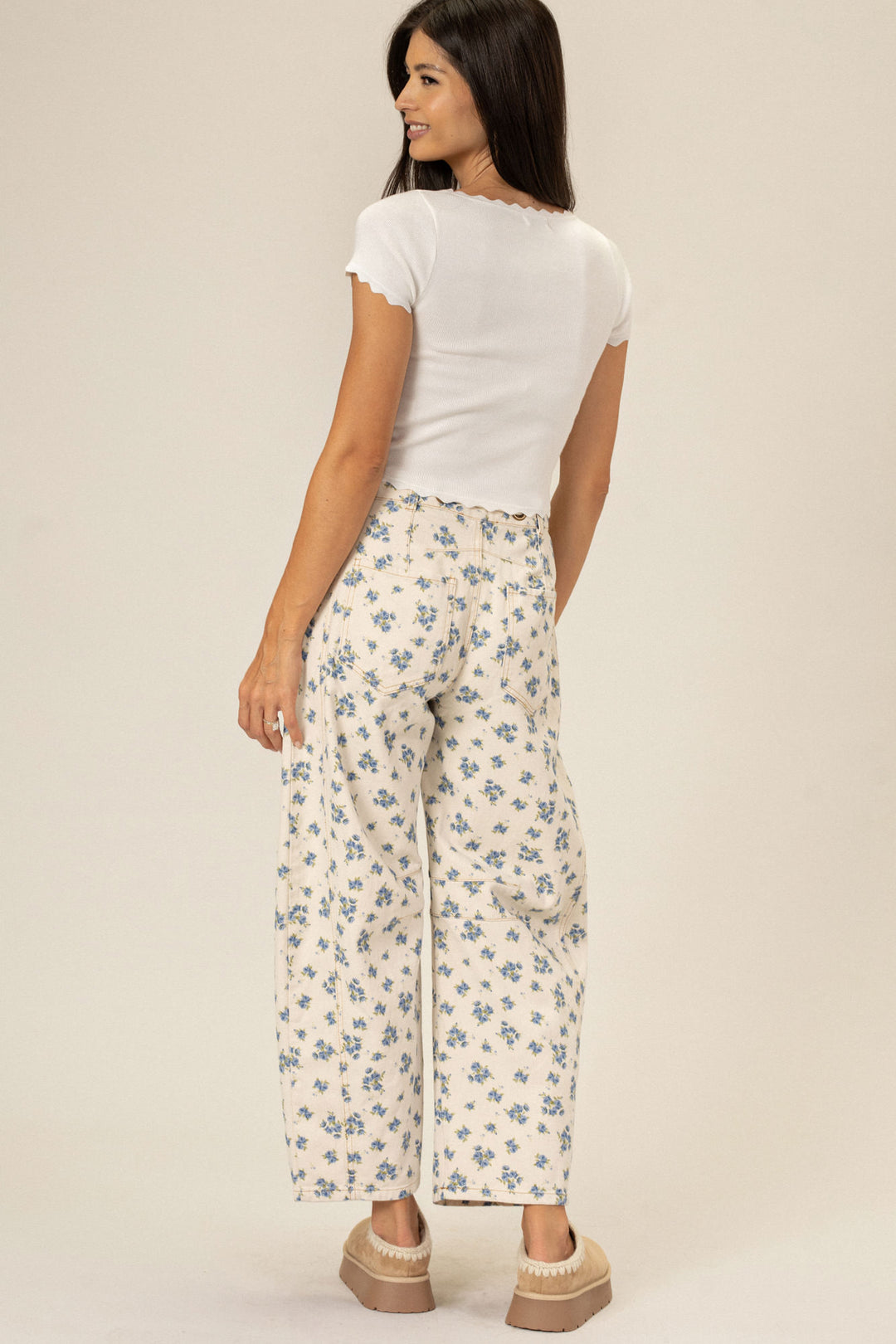 A young woman in white shirt and floral printed wide-leg pants, posing with hands on hips and smiling.