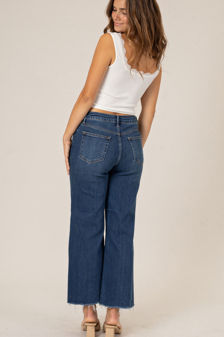 A young woman wearing a white lace-trimmed tank top and wide-leg blue jeans, standing against a beige background.