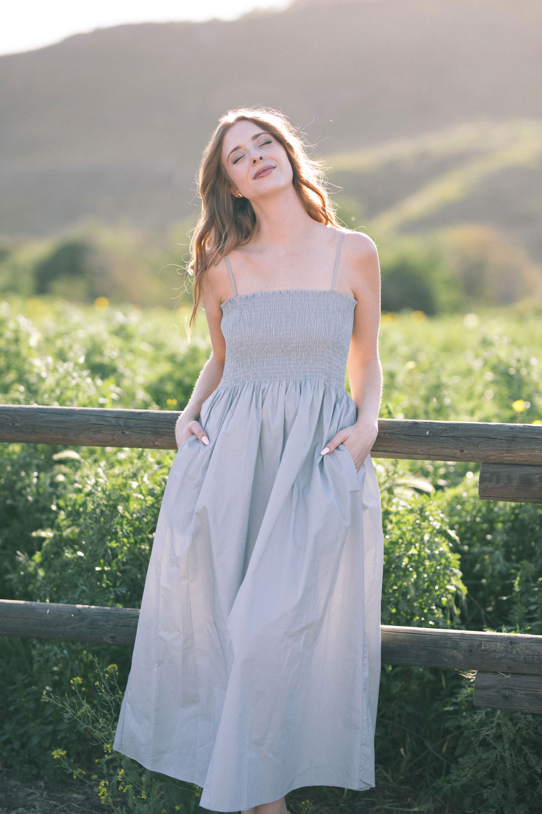 A long, gray maxi dress with a smocked bodice and spaghetti straps, set against a backdrop of lush greenery.