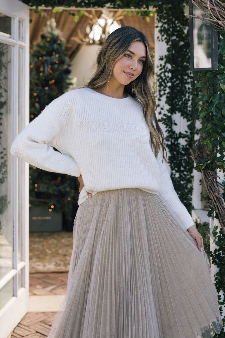 A woman wearing a white, ribbed knit sweater with "Champion" embroidered in script, paired with a beige pleated midi skirt, standing in a garden setting.