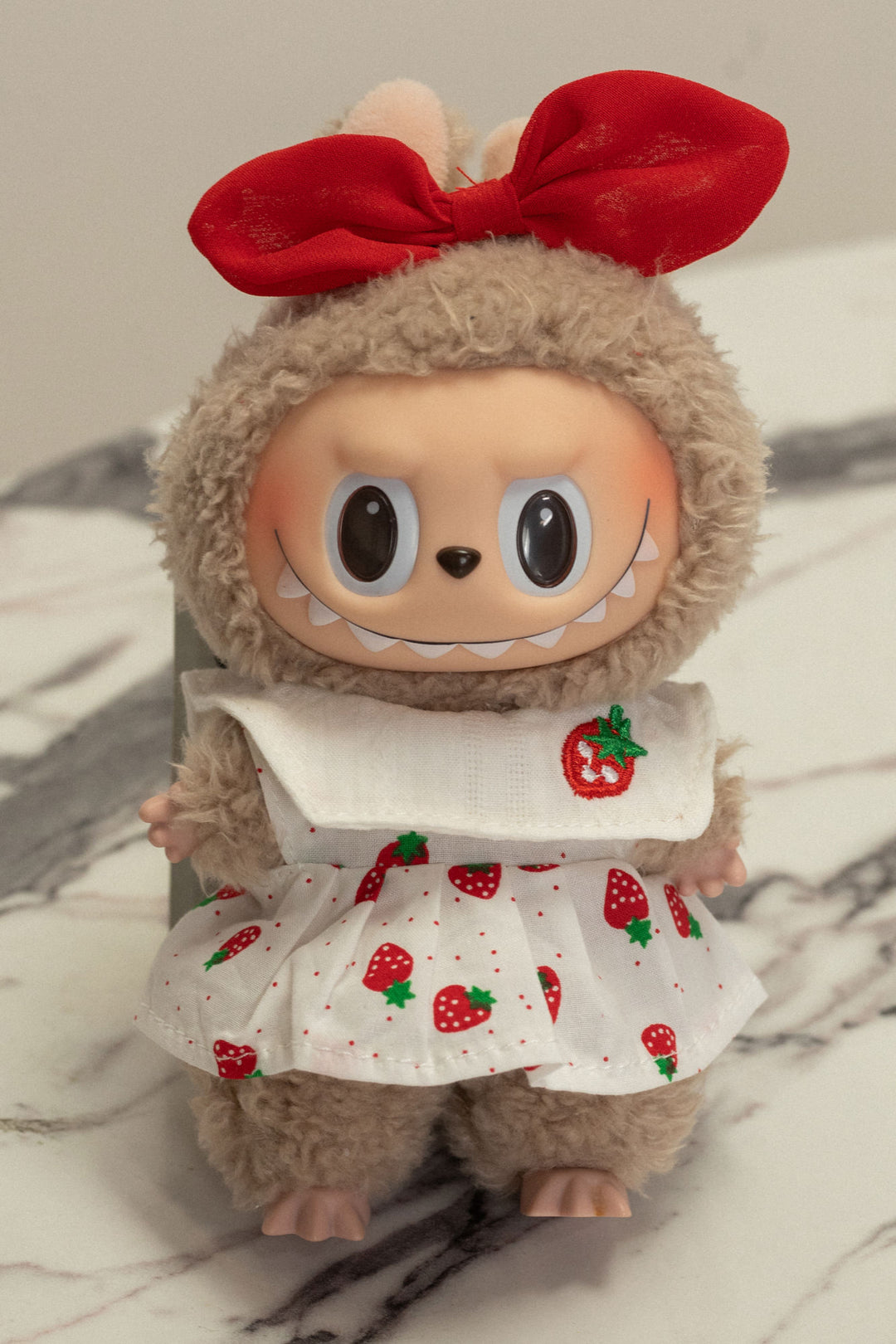 Plush gray stuffed animal with large eyes, wearing a red bow and a white dress with a strawberry print.