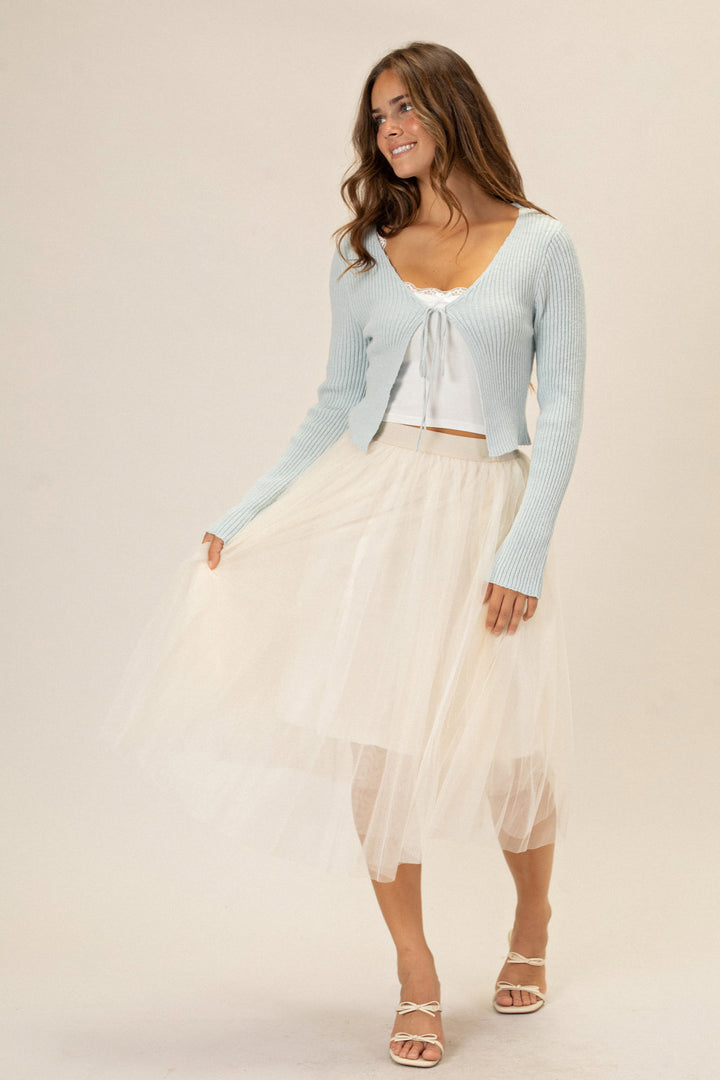 A smiling woman wearing a mint green ribbed knit sweater with a front tie detail, paired with a flowing white tulle midi skirt, and strappy beige sandals.