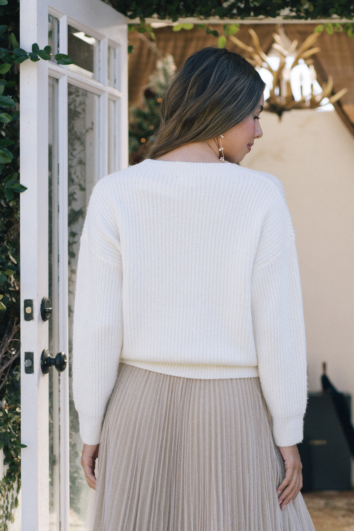 A white knitted pullover sweater with a ribbed texture, worn with a pleated beige skirt, shown from the back.