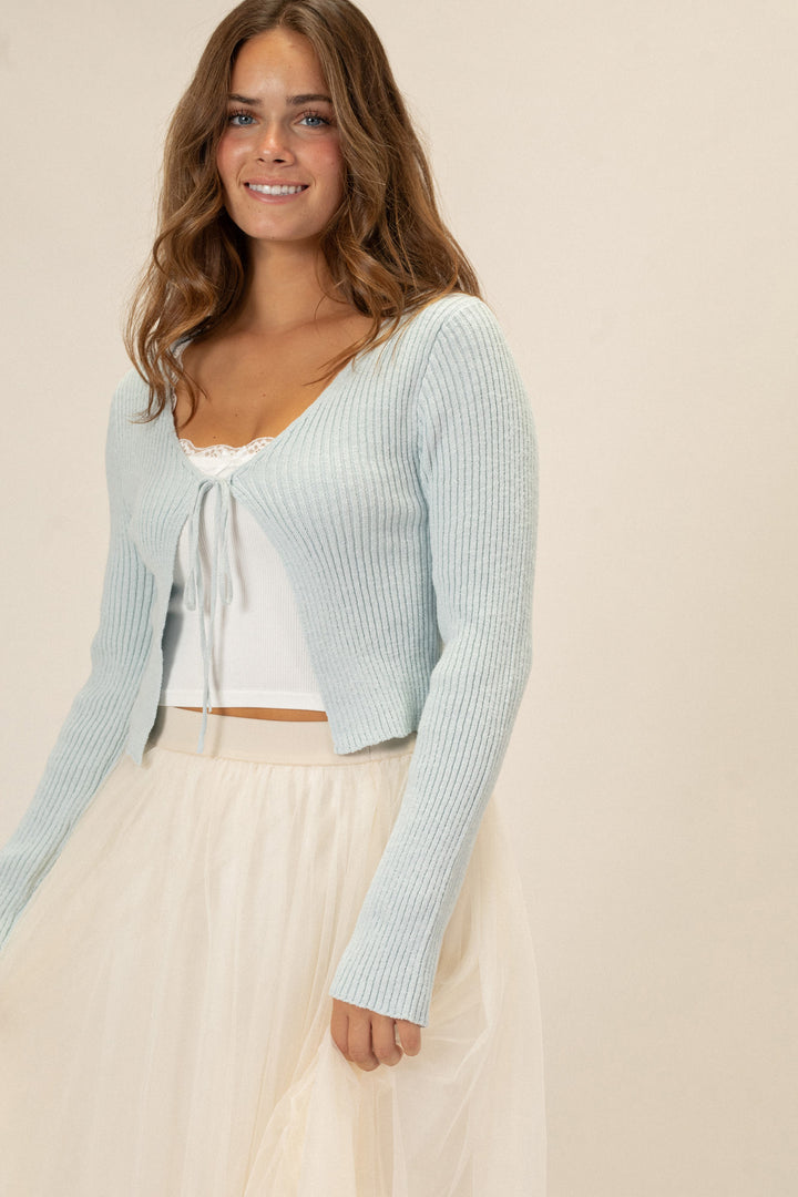 A pale blue ribbed knit top with a front tie detail, modeled by a young woman with wavy brown hair.