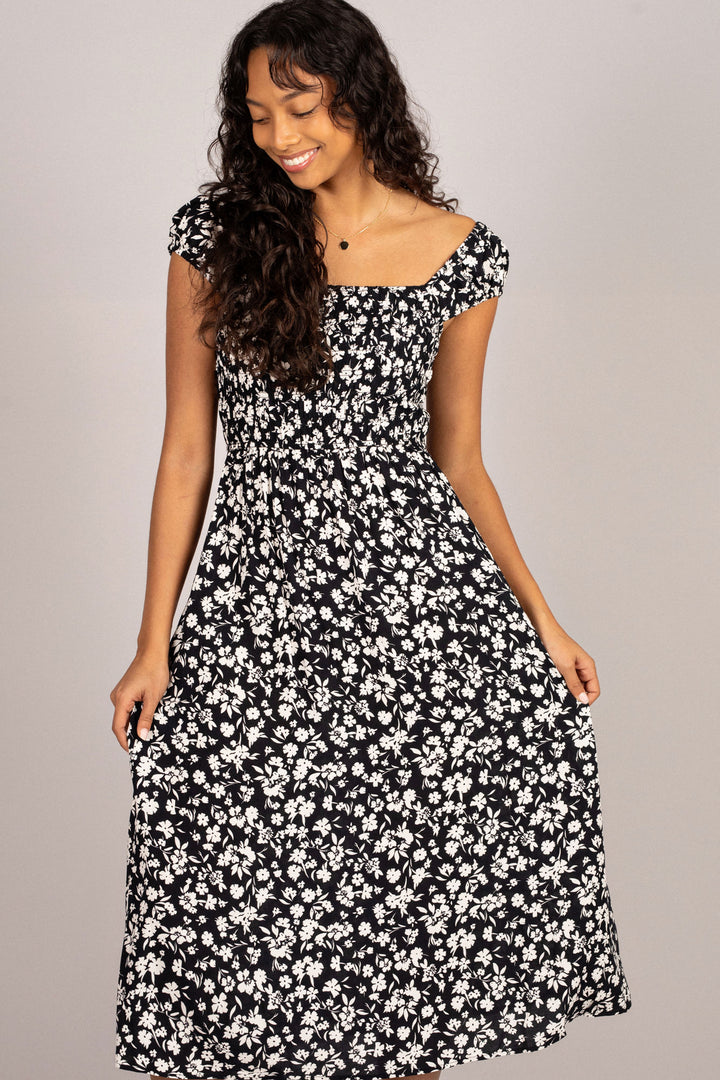 A black dress with a floral print, fitted bodice, and flared midi skirt, modeled by a smiling woman with curly dark hair.