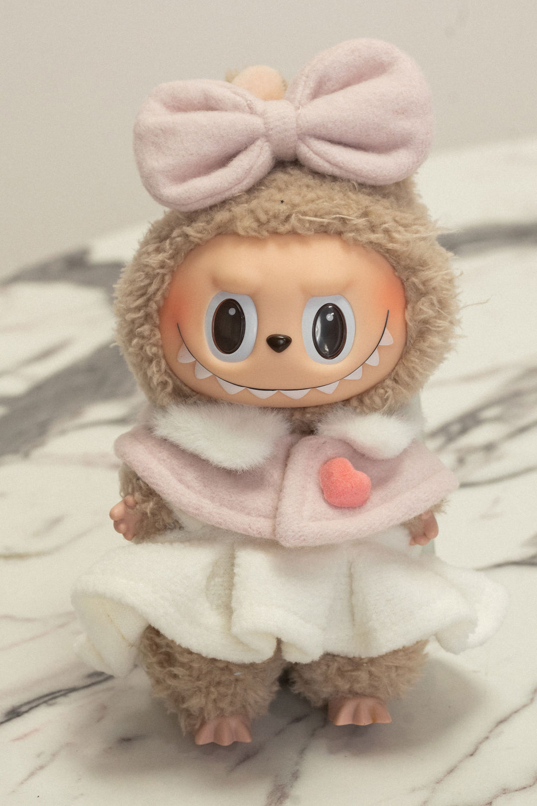 Furry gray plush toy with large eyes, bow headband, and fuzzy pink vest with heart detail.