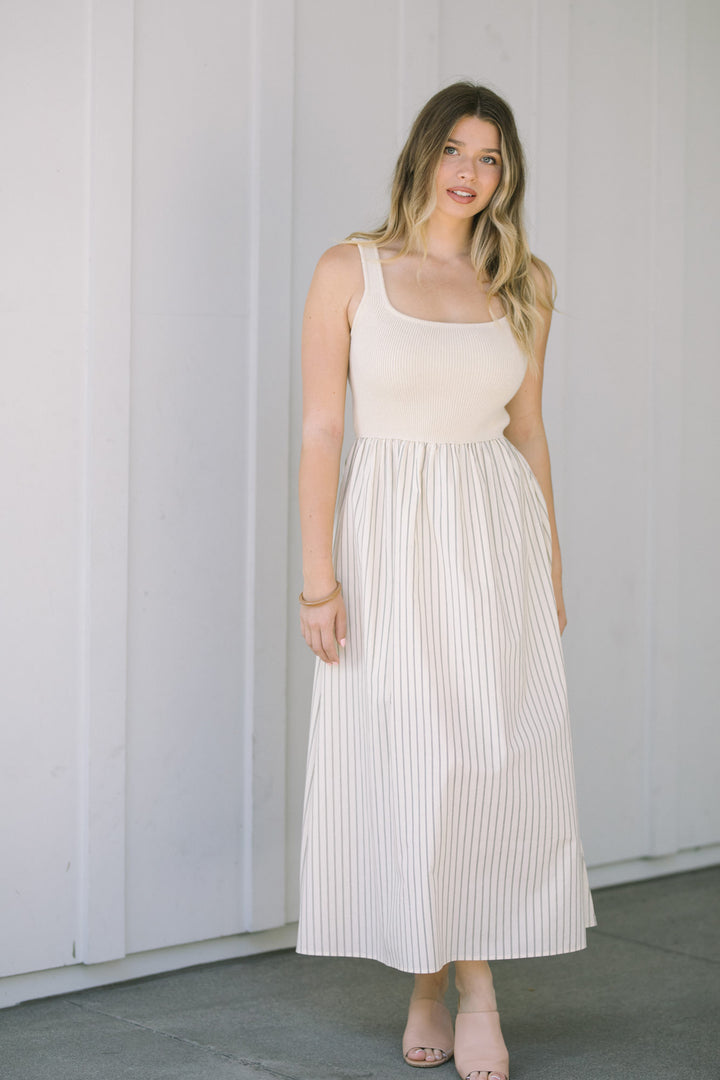 A young woman wearing a beige knit tank top and a long pleated striped skirt, posing against a white wall.