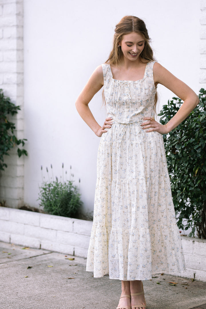 A young woman smiling and posing in a white floral print dress with a tiered, ruffled skirt and adjustable waist.