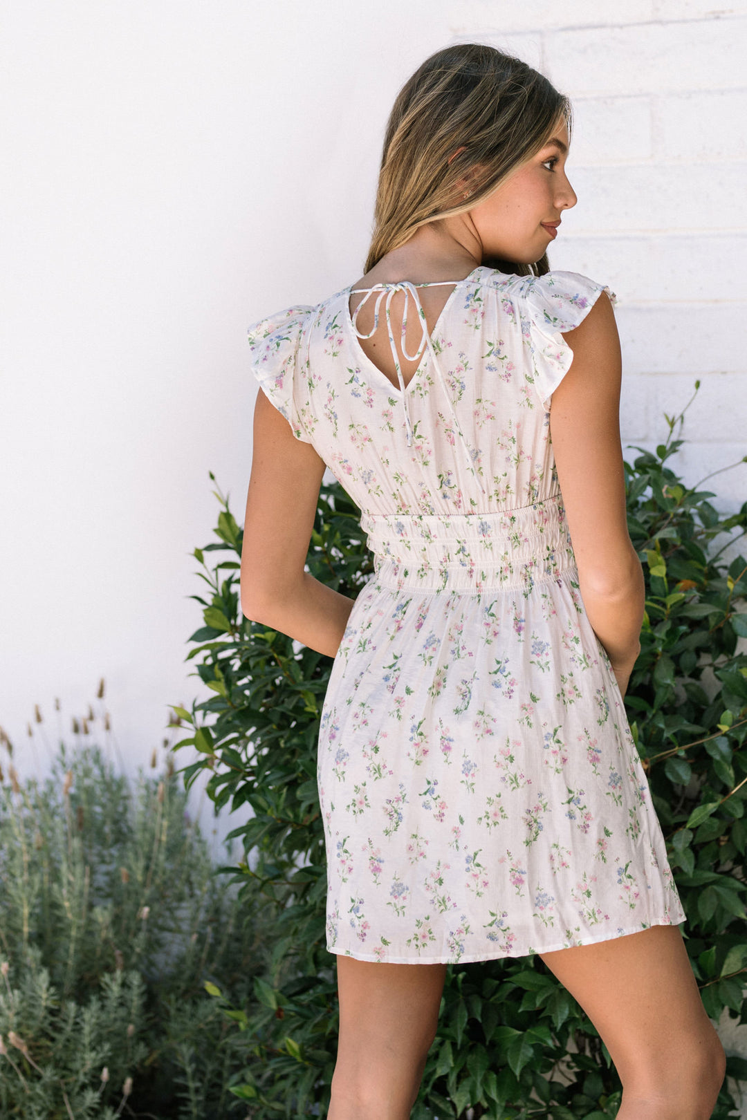 A floral-printed white mini dress with ruffled cap sleeves, a smocked waist, and a tie-back detail.