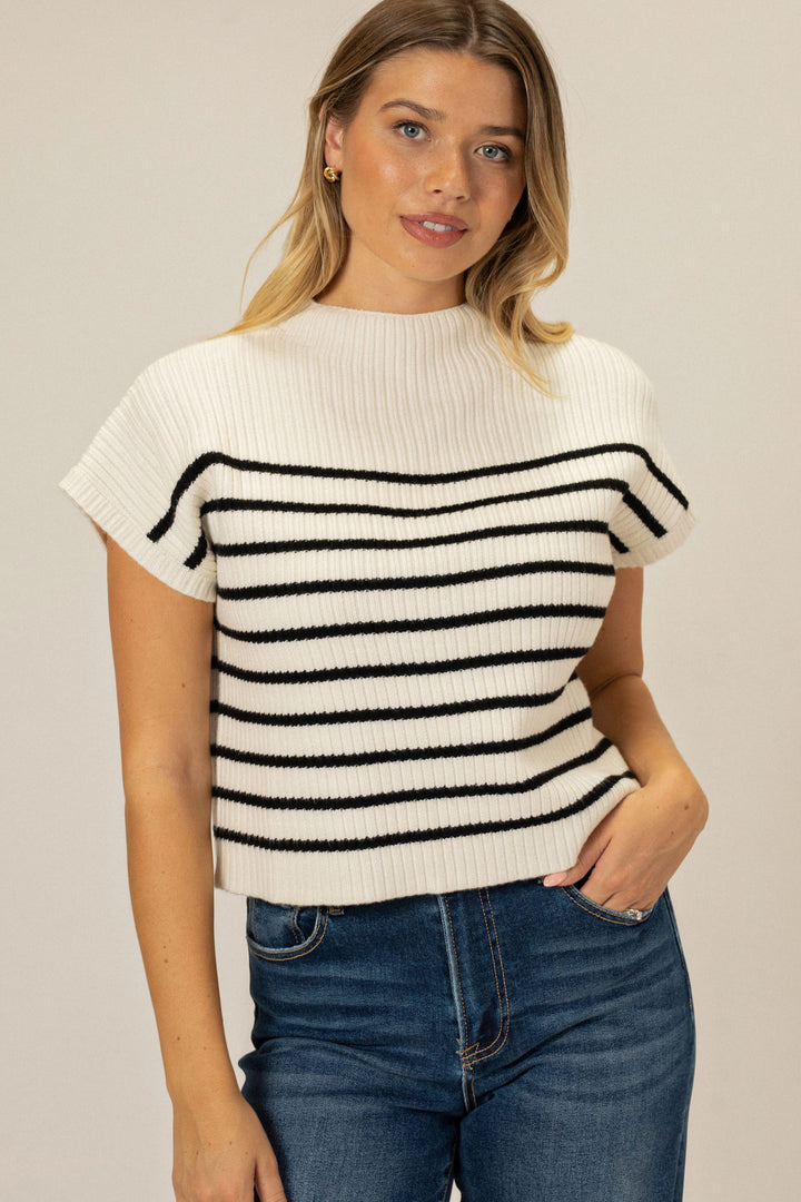 A white and black striped mock-neck knit top with short sleeves, featured on a young woman with long blonde hair.