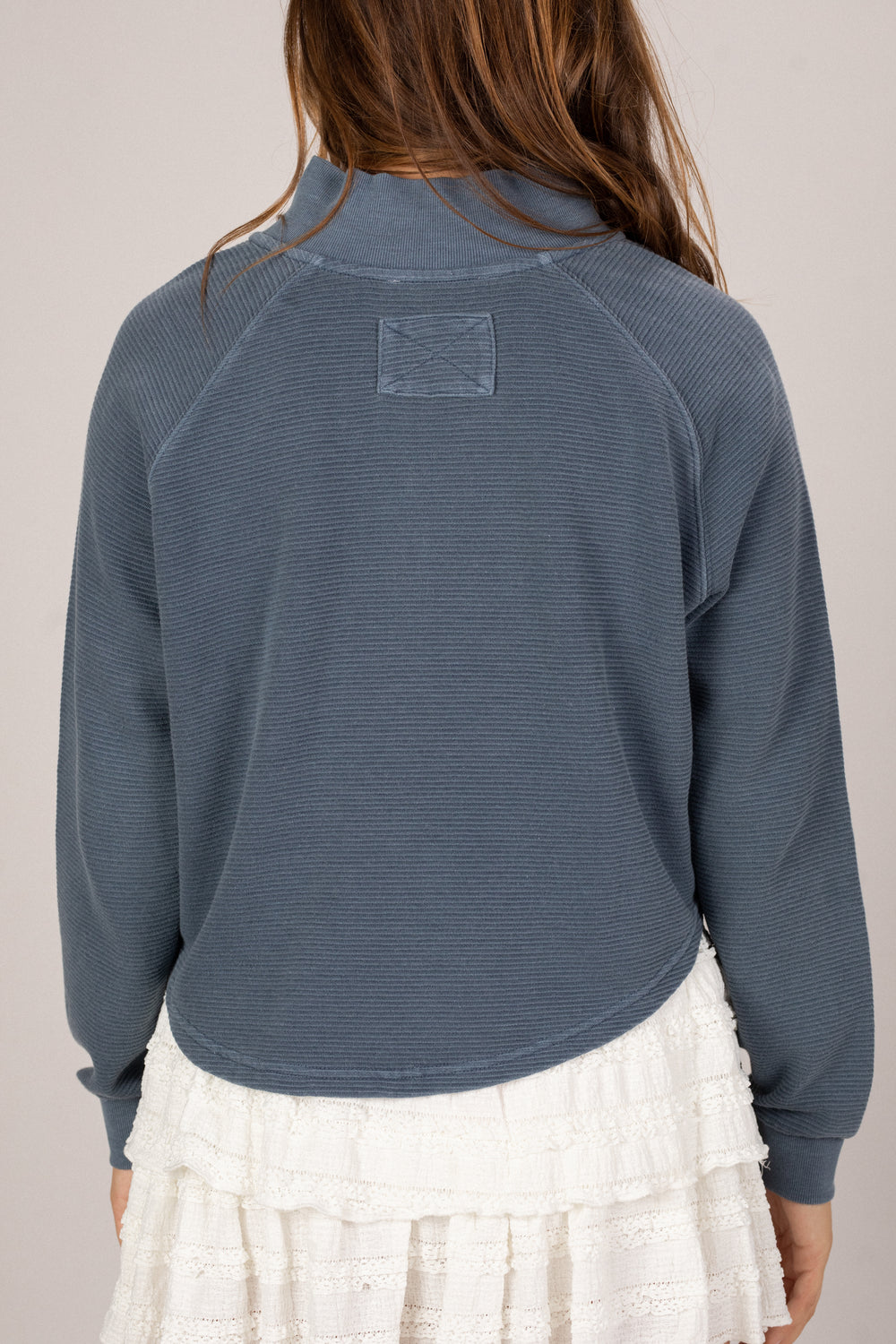 Person wearing a blue textured sweater with a white ruffled skirt on a neutral background facing the back.