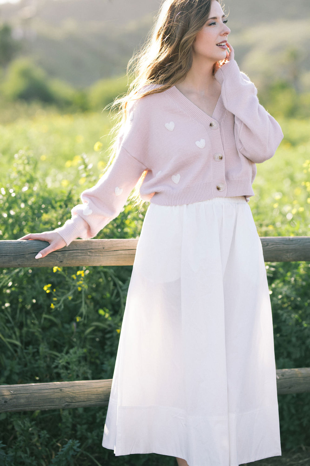 A soft pink knitted cardigan with white heart-shaped buttons, featuring a V-neckline and relaxed silhouette, creating a cozy and feminine look.