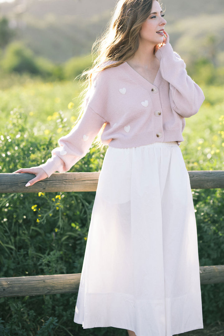 A soft pink knitted cardigan with white heart-shaped buttons, featuring a V-neckline and relaxed silhouette, creating a cozy and feminine look.