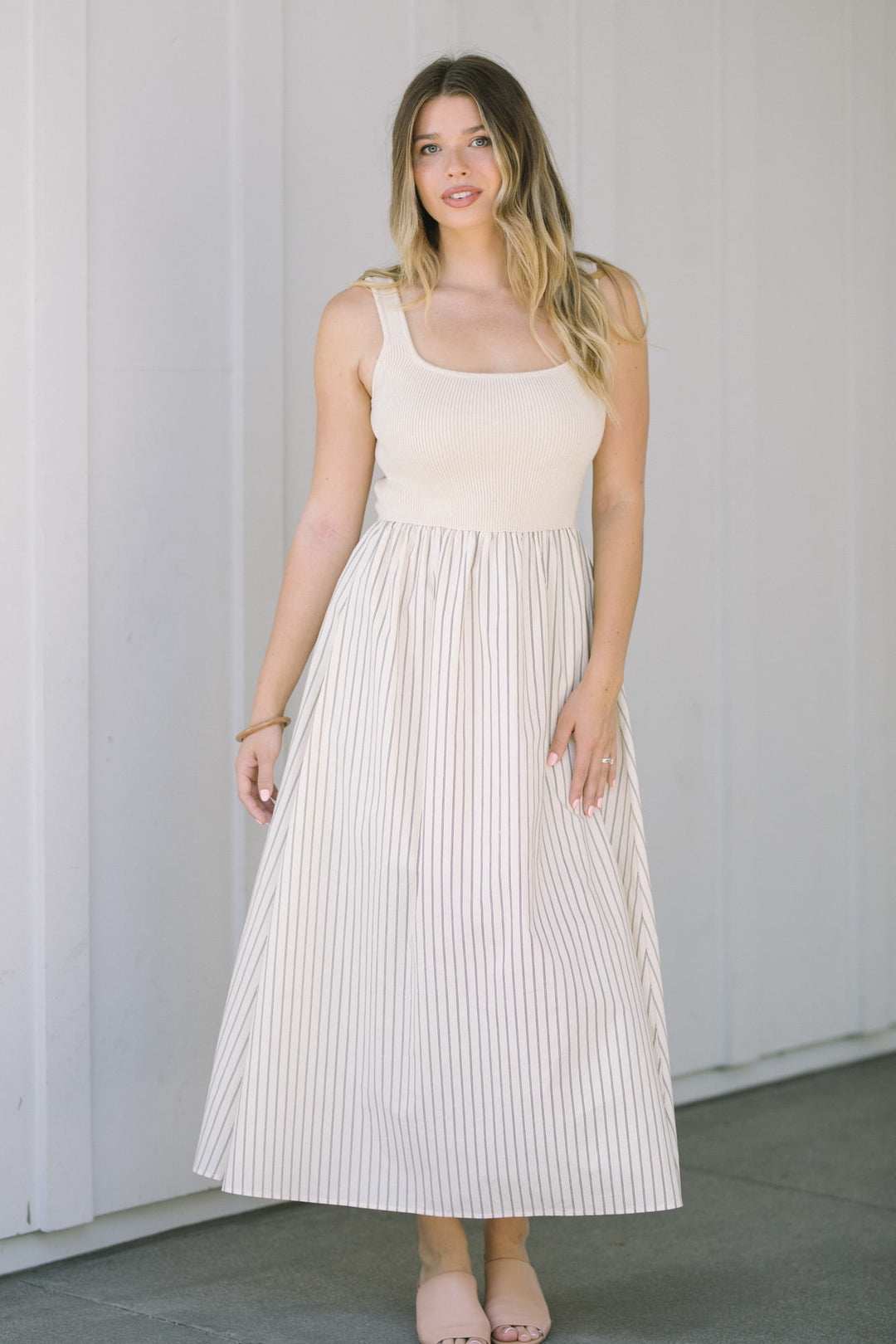A sleeveless knee-length dress with a fitted bodice and a pleated, striped skirt in a neutral beige and white color scheme.