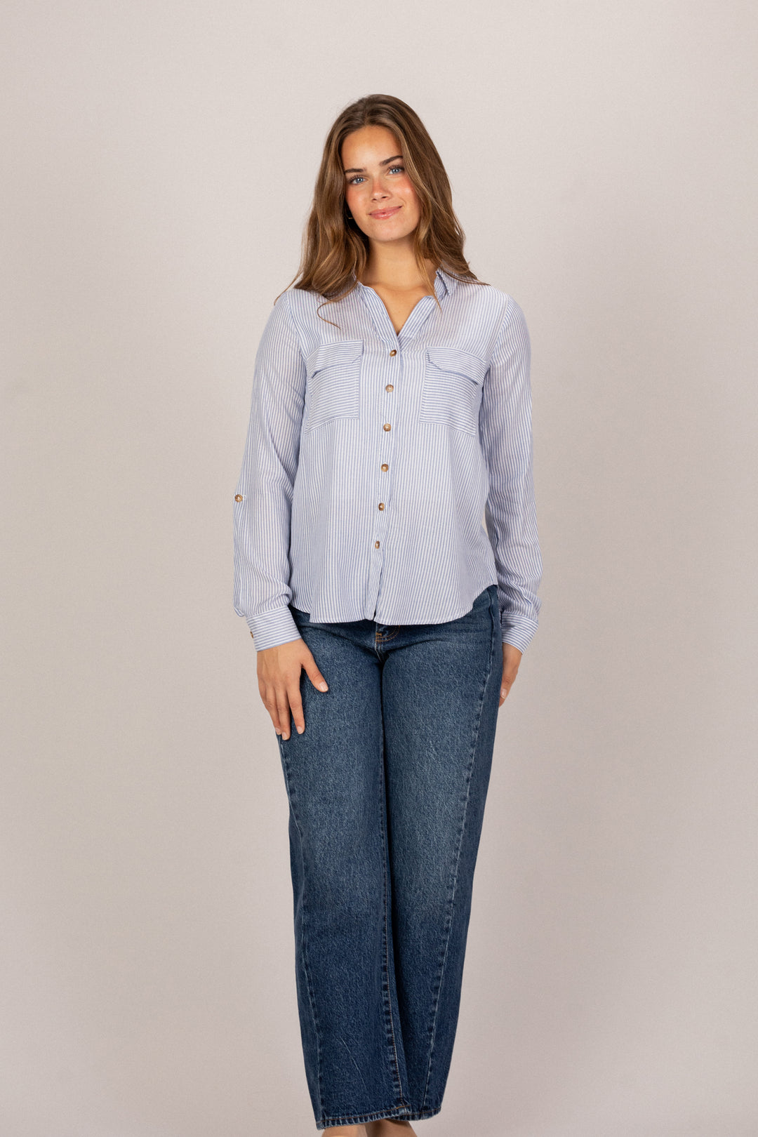 A young woman wearing a striped button-up shirt and blue jeans, standing against a neutral background.