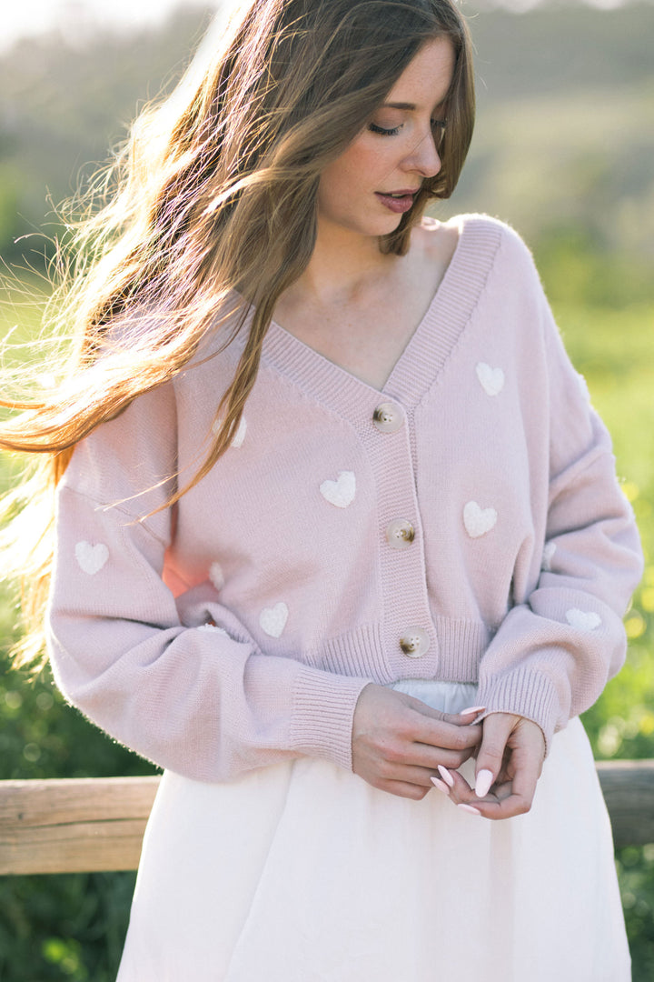 A soft, light pink cardigan with white heart motifs, featuring a V-neck and button-up design, perfect for layering over outfits.