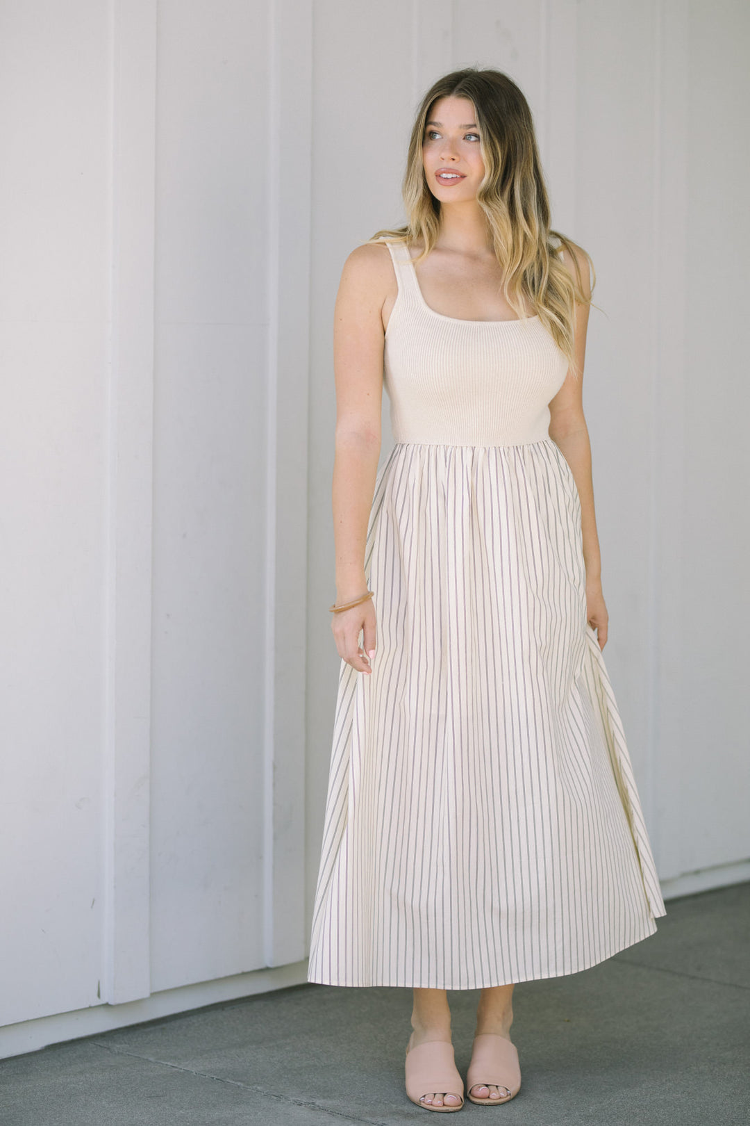 A woman wearing a sleeveless, knee-length dress with a beige knit bodice and a striped pleated skirt, styled with nude sandals.