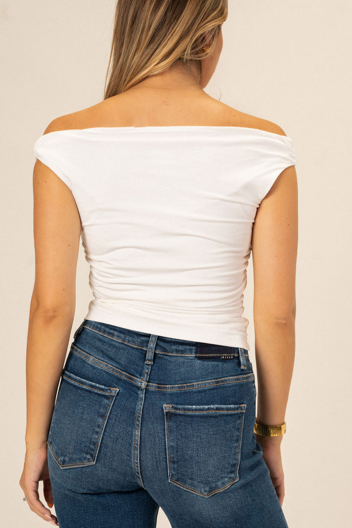 A white off-the-shoulder top with a fitted, ruched bodice, worn by a model with long, golden-brown hair.