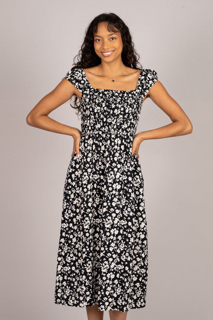 A young woman wearing a black floral midi dress with a smocked bodice, puff sleeves, and a flowing skirt, standing against a neutral background.