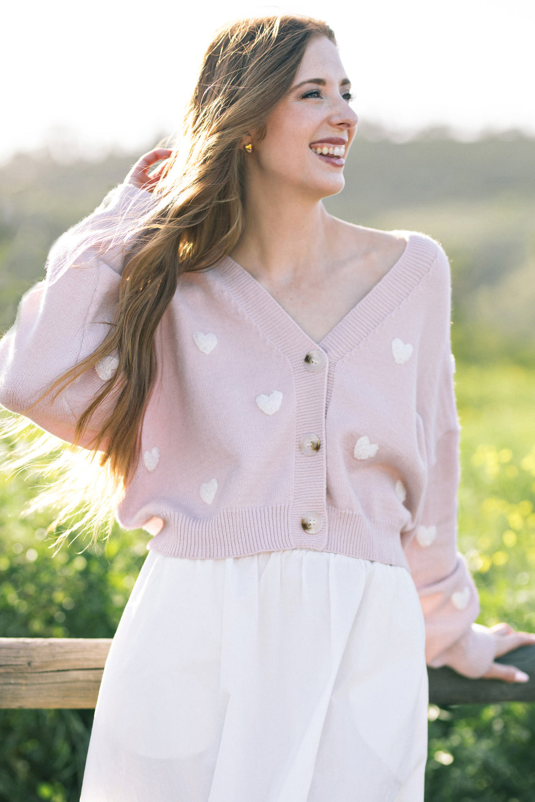 A smiling woman wearing a pink knit cardigan with white heart patterns, standing in a lush outdoor setting.