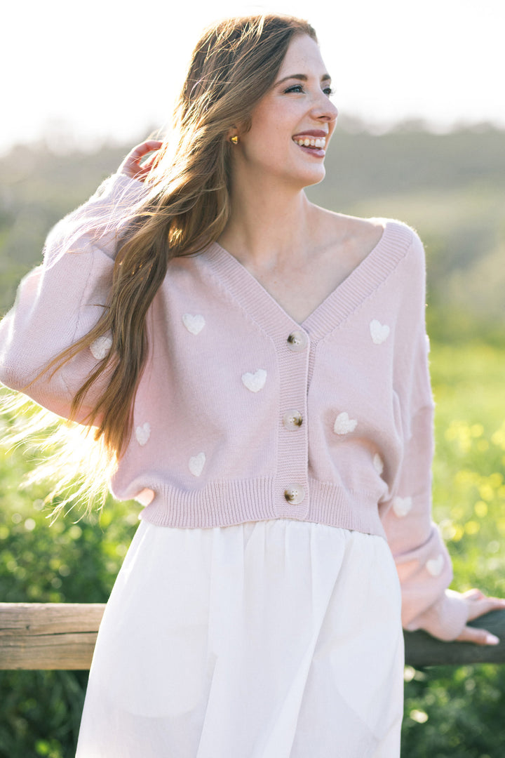 A smiling woman wearing a pink knit cardigan with white heart patterns, standing in a lush outdoor setting.