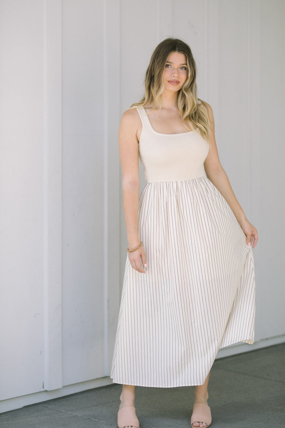 A woman wearing a cream-colored sleeveless dress with a pleated skirt featuring thin vertical stripes, standing in front of a gray wall.