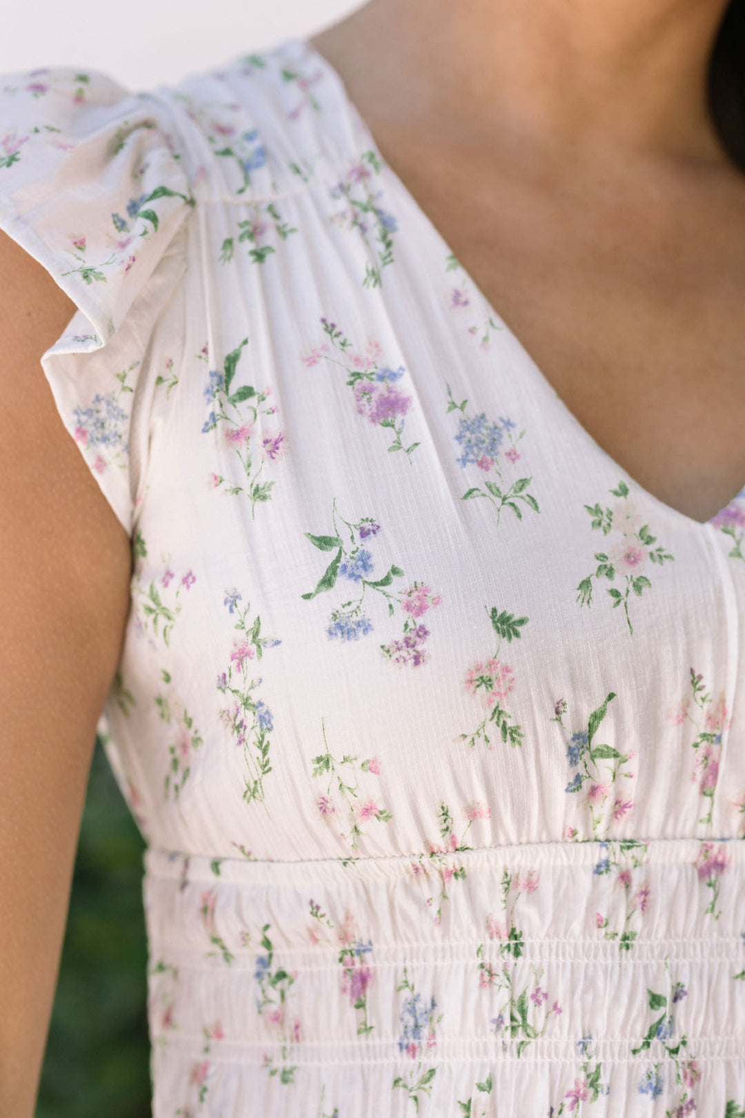 A white dress with a floral print, featuring a pleated, shirred waist and a flowing, ankle-length silhouette.