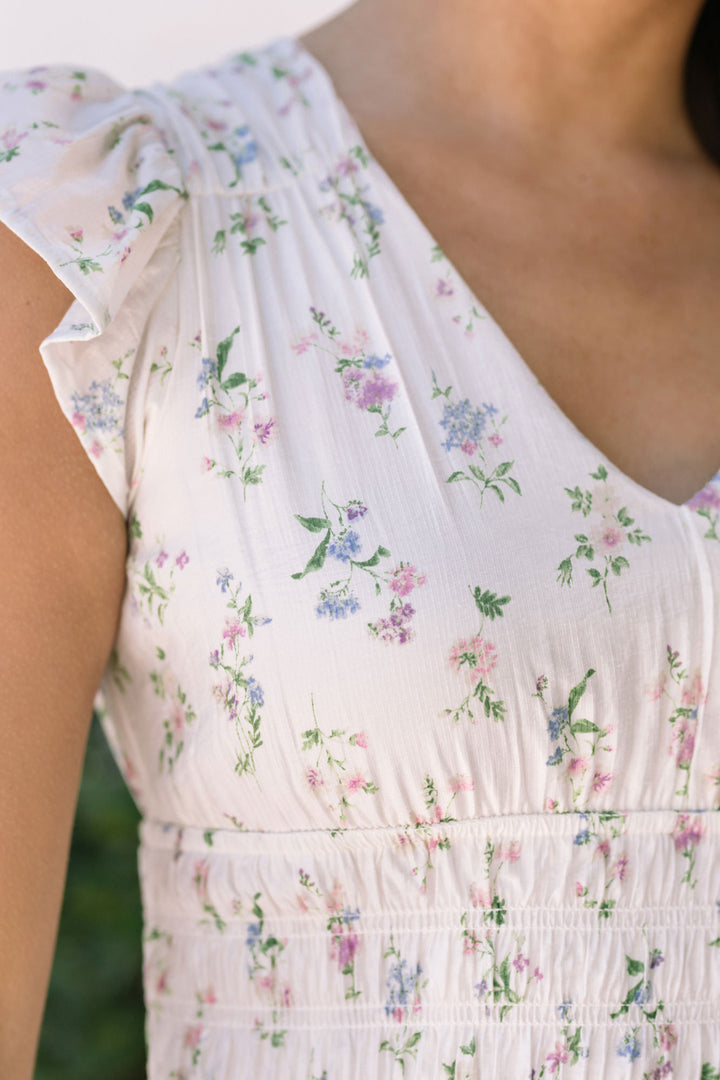A white dress with a floral print, featuring a pleated, shirred waist and a flowing, ankle-length silhouette.