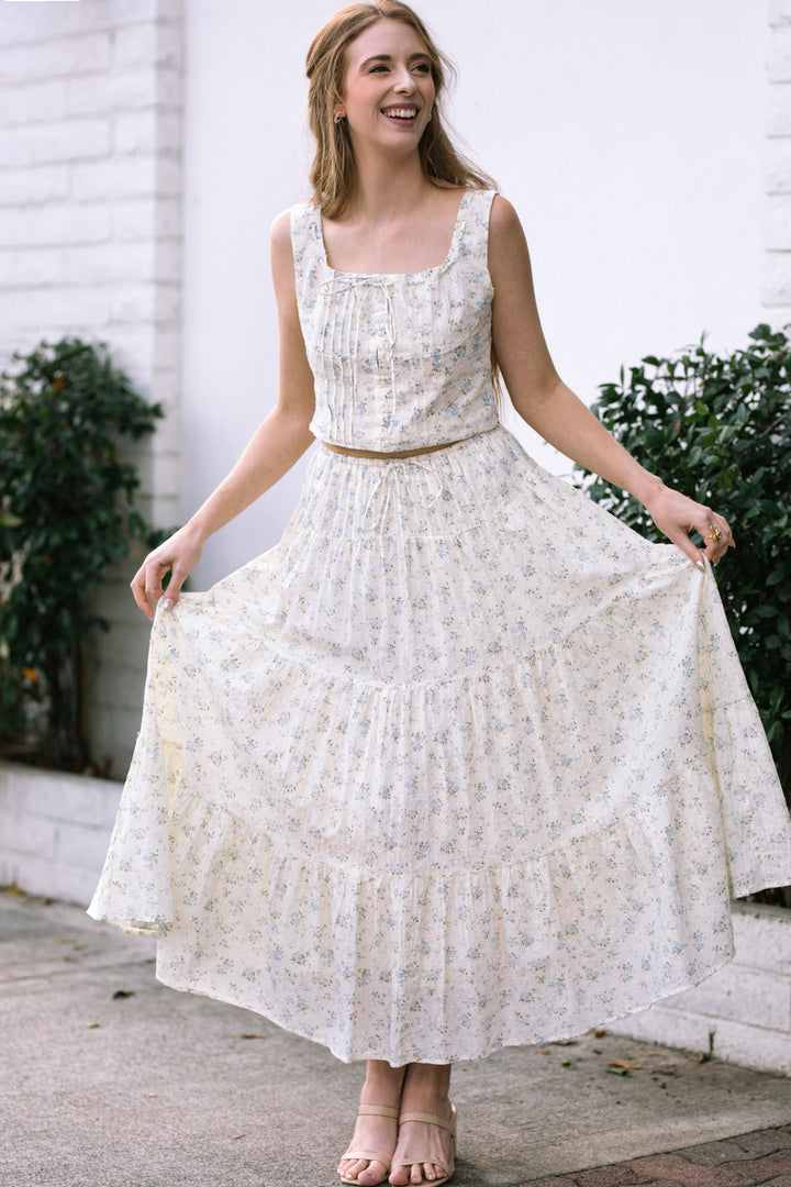 A white, floral-patterned sundress with a pleated bodice, full skirt, and adjustable waistband that flows gracefully.