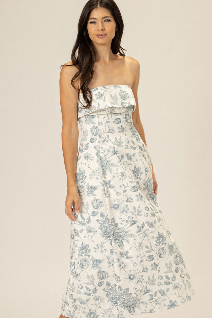 A strapless, floral maxi dress in a white and gray color palette, featuring a ruffled bust and a flowing, floor-length skirt.
