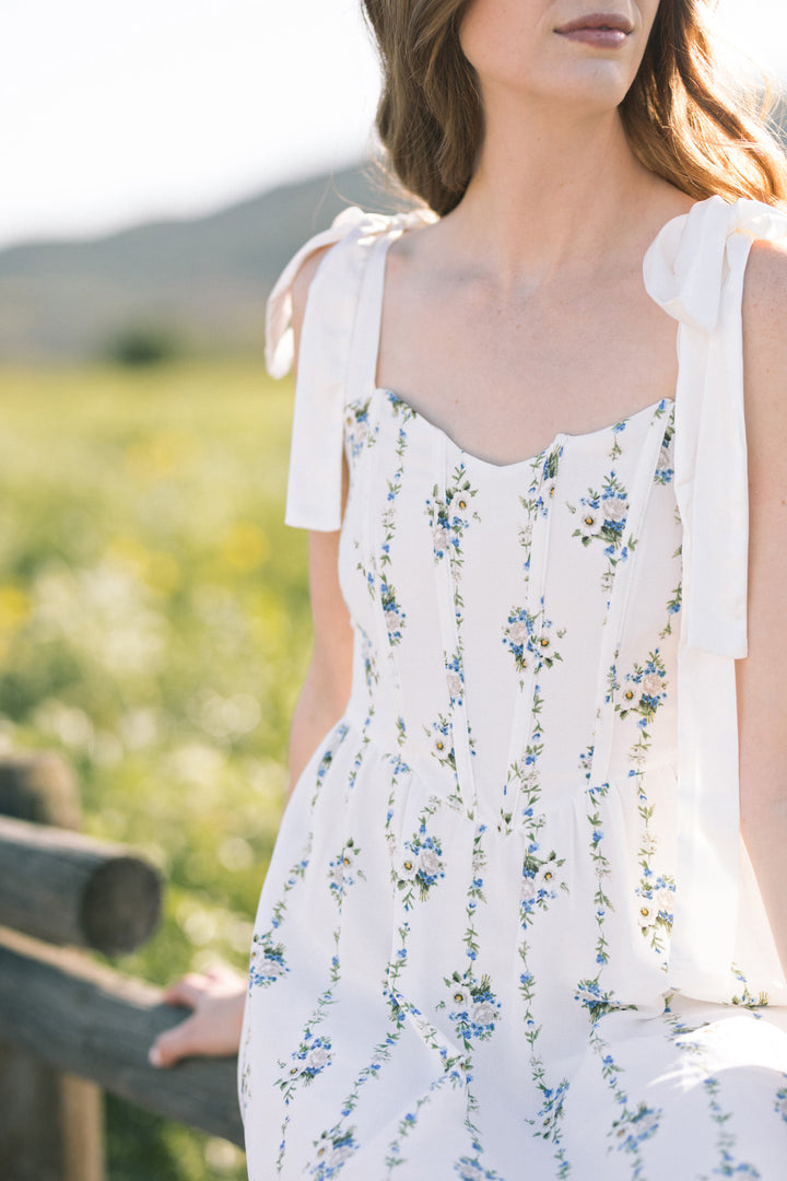 A white, floral-print dress with ruffled shoulder straps, styled with a fitted bodice and flowing skirt, against a scenic outdoor backdrop.