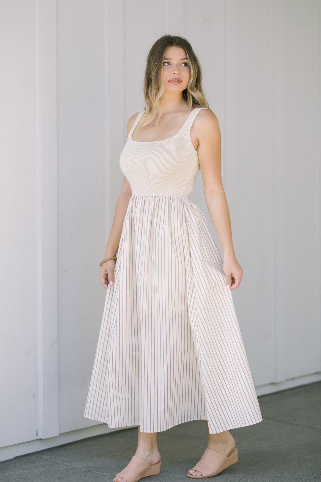 A young woman wearing a sleeveless, knee-length dress with a scoop neckline and a pleated, striped skirt in beige and white.