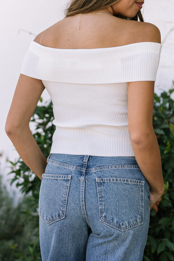 A white off-the-shoulder ribbed knit top with a delicate chain necklace, modeled by a woman with long brown hair against a floral background.