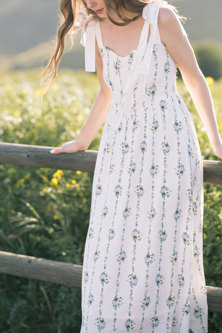 A white maxi dress with a floral pattern, featuring spaghetti straps and a high waistline, set against a natural, scenic backdrop.