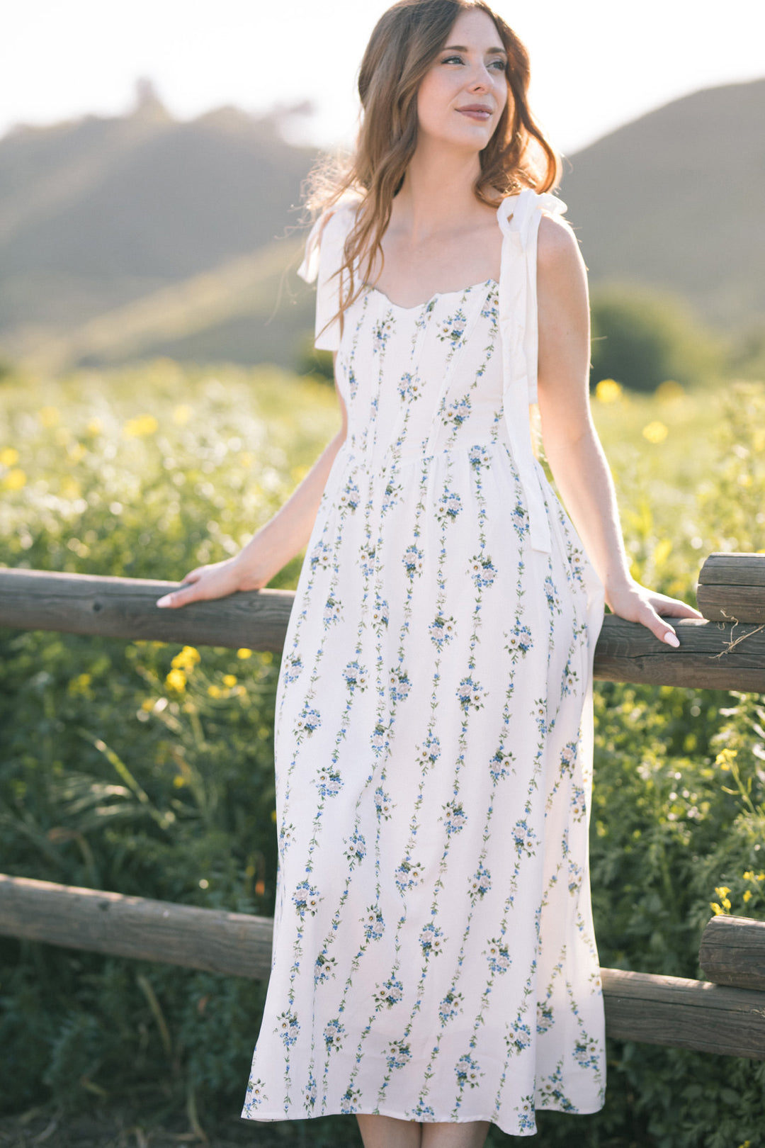 A young woman wearing a white floral maxi dress with spaghetti straps, standing in a field of wildflowers against a mountain backdrop.