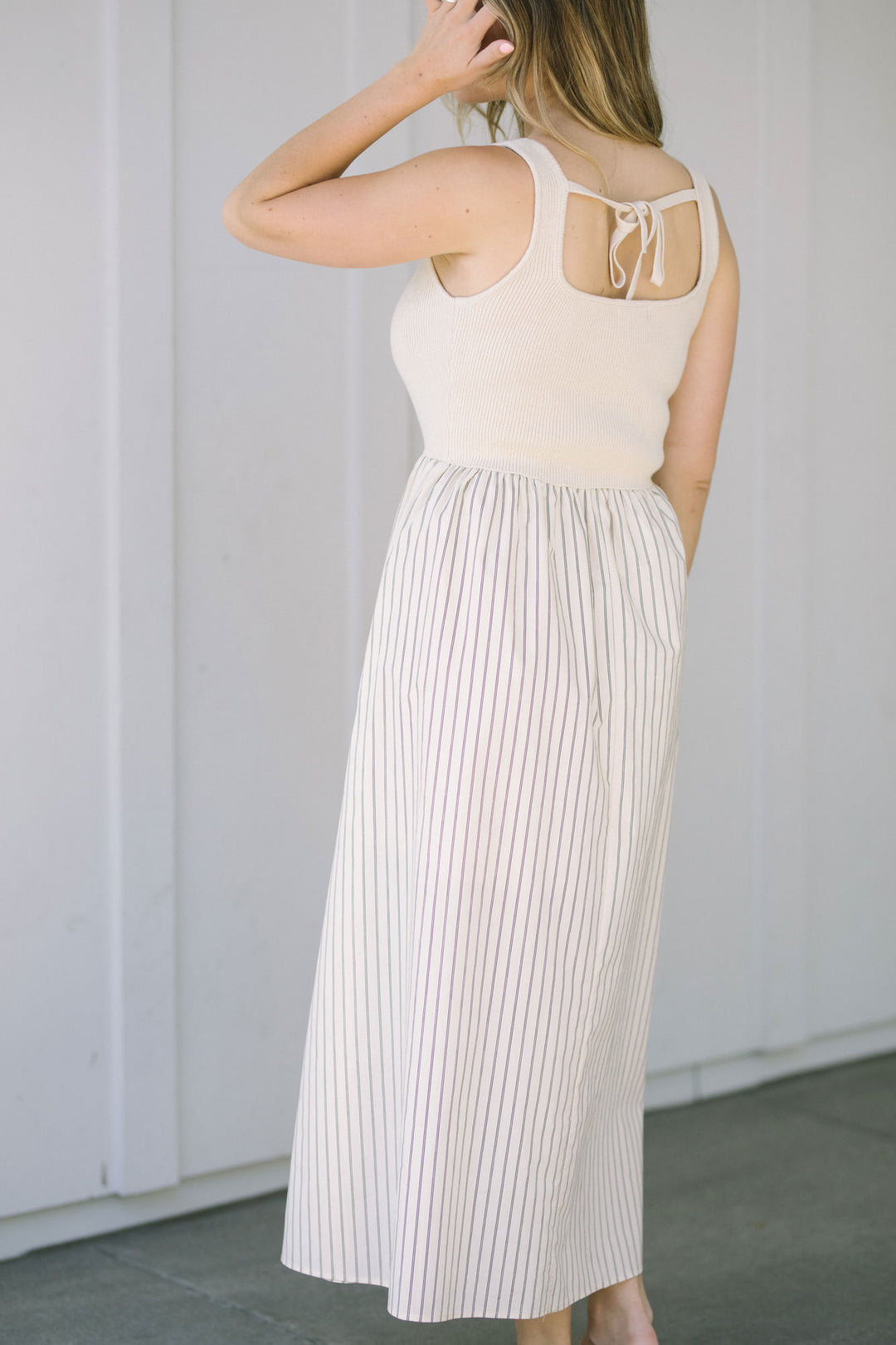 Image shows a cream-colored knit top with a strappy back detail, paired with a long white and pink striped skirt. The skirt features a high waist and a flowing, pleated silhouette.