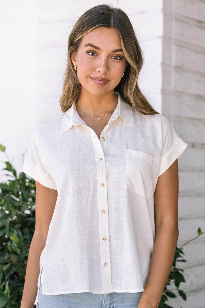 A young woman wearing a white button-down short-sleeve shirt, with her hair styled in waves and a simple necklace. She is standing in front of a white brick wall.