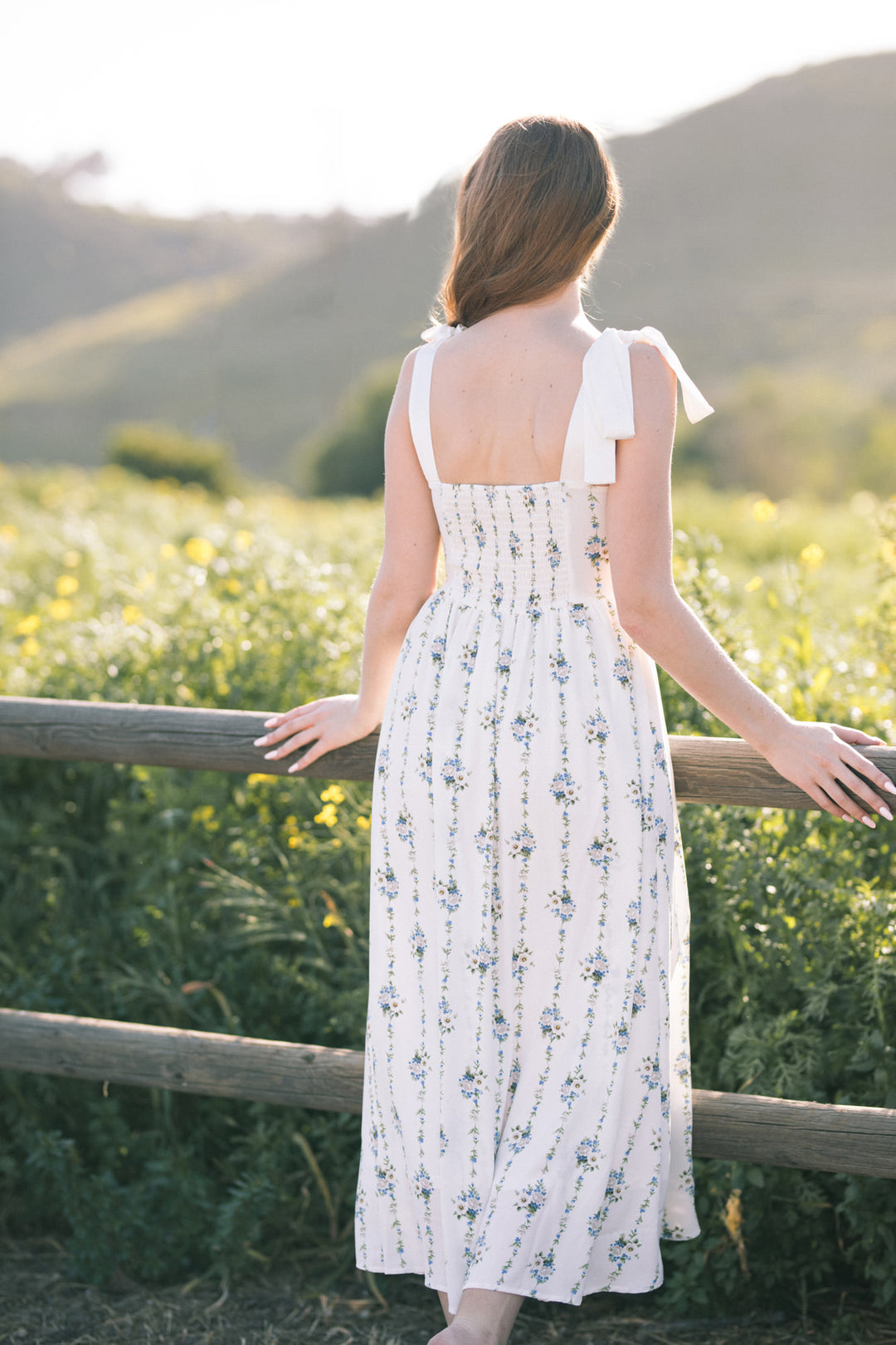 A woman wearing a white sleeveless dress with blue floral print, standing in a field with mountains in the background.
