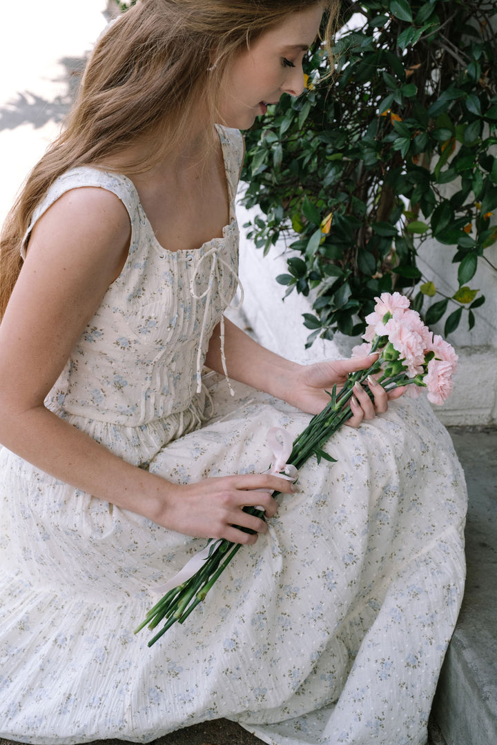 A woman in a white and blue floral print dress holds a bouquet of pink peonies, surrounded by greenery.