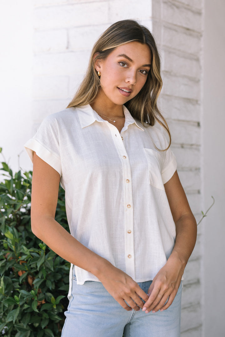 A young woman with long, wavy brown hair models a white button-down shirt with short sleeves. The shirt features a classic collar and front button closure, creating a relaxed, casual style.