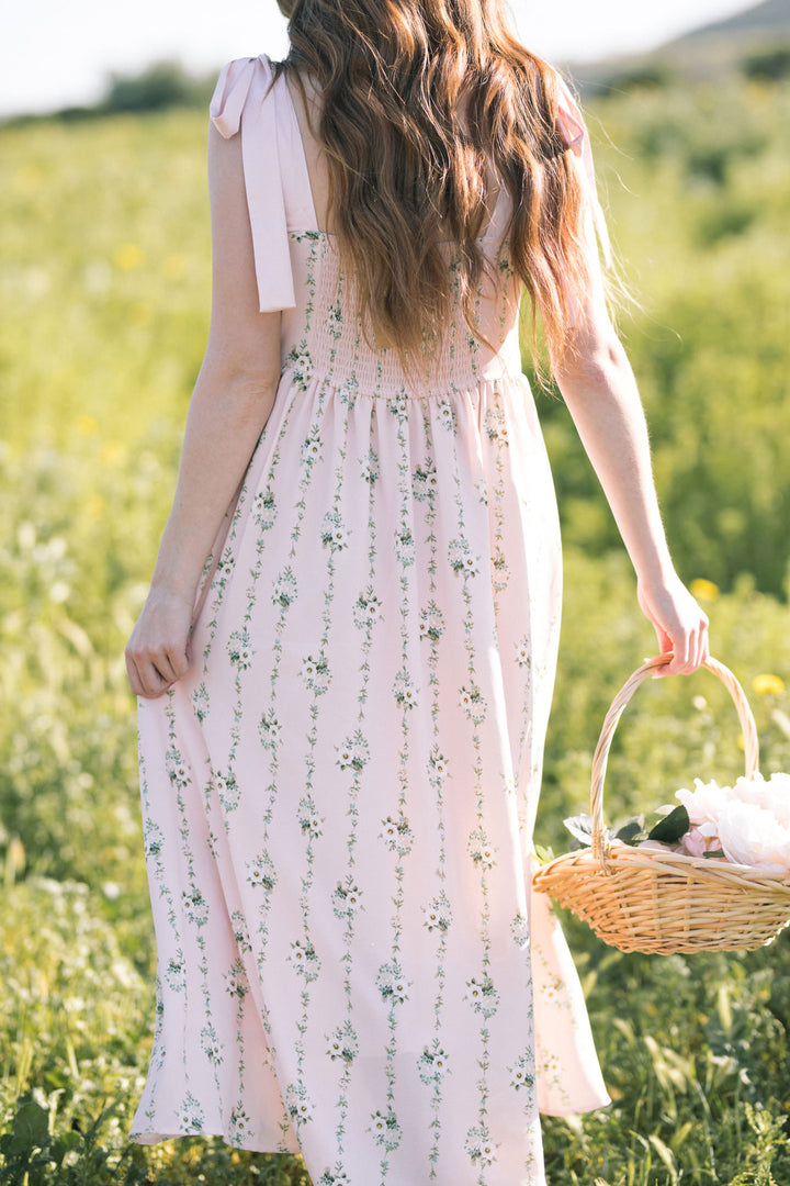 A pink floral maxi dress with puffed sleeves, a fitted bodice, and a flowing, ankle-length skirt, held in the model's hand is a woven wicker basket filled with white flowers.