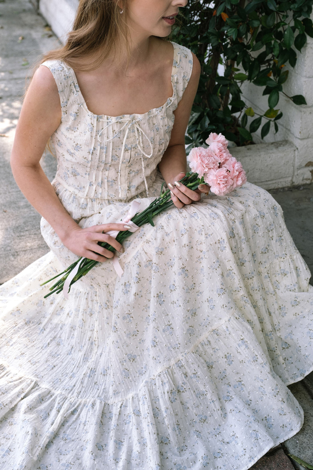 A young woman in a flowing white dress with blue floral print, holding a bouquet of pink peonies, standing against a background of lush greenery.