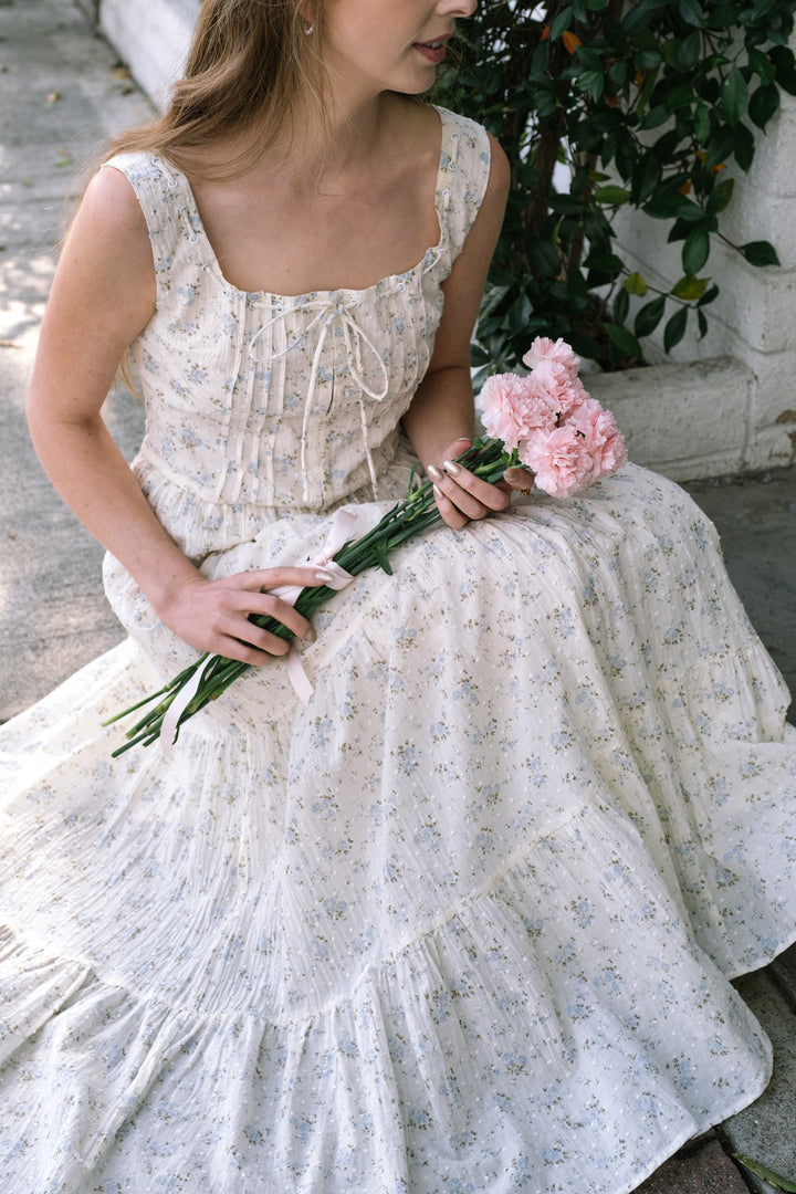 A young woman in a flowing white dress with blue floral print, holding a bouquet of pink peonies, standing against a background of lush greenery.