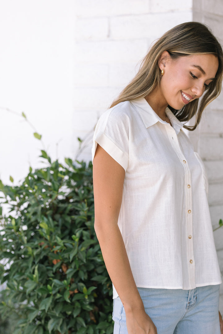A young woman in a white button-down shirt smiling against a backdrop of lush greenery.