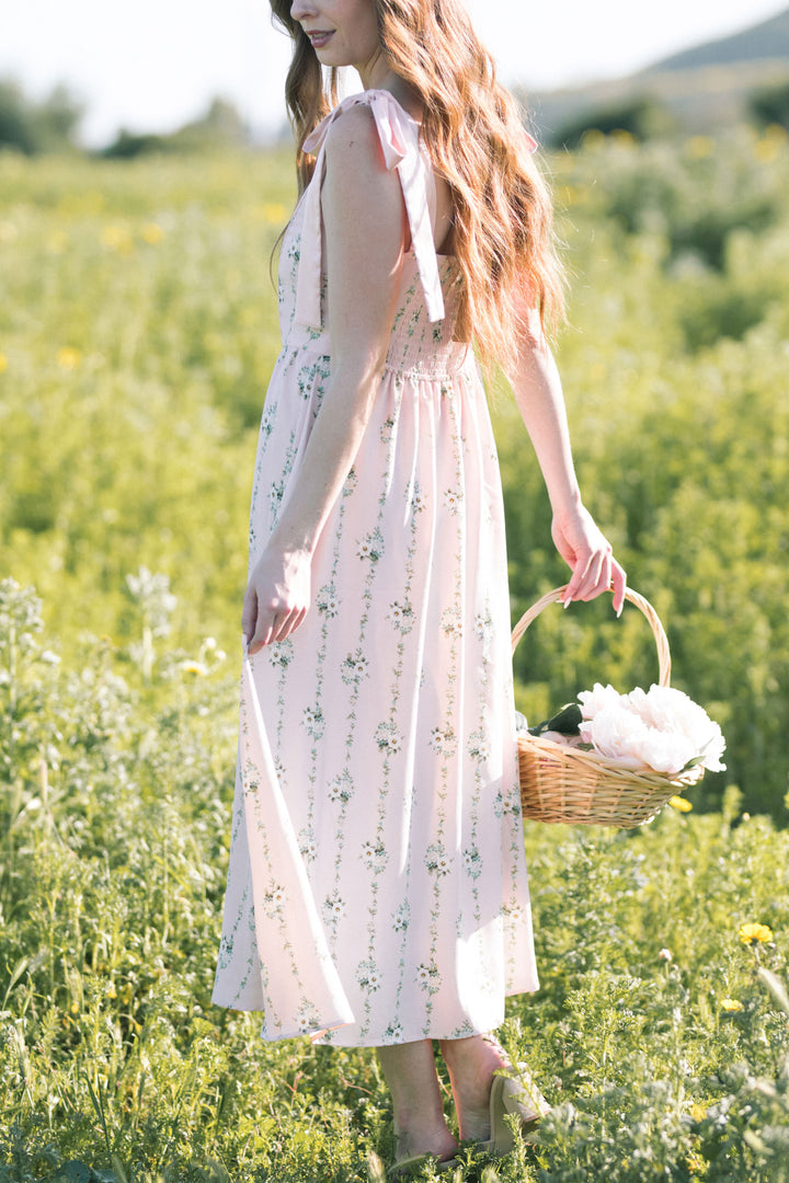 A young woman wearing a long, floral print dress with puff sleeves and a flowing skirt, holding a basket of white flowers while standing in a field of yellow flowers.