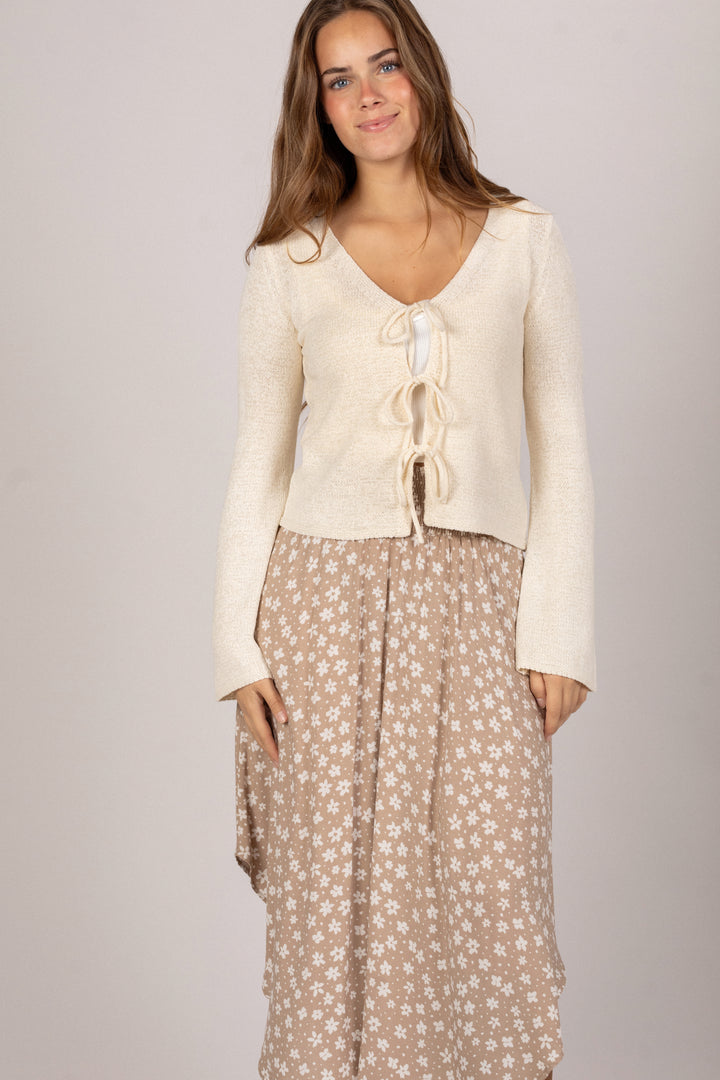 A cream-colored knit top with a front tie detail, paired with a beige floral-patterned skirt, worn by a model with long brown hair.