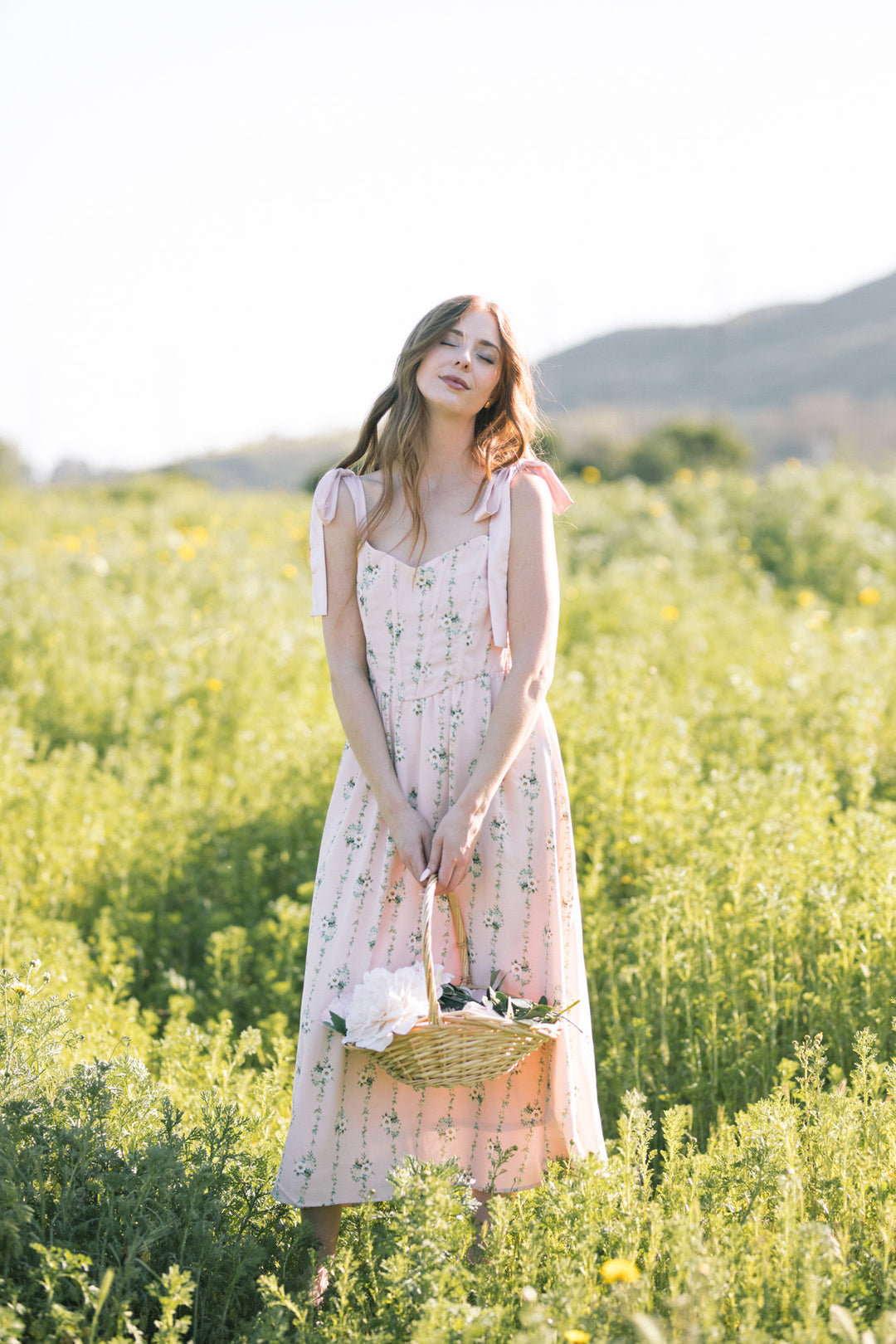 A woman in a long floral print dress stands in a field of yellow flowers, holding a wicker basket, with mountains in the background.