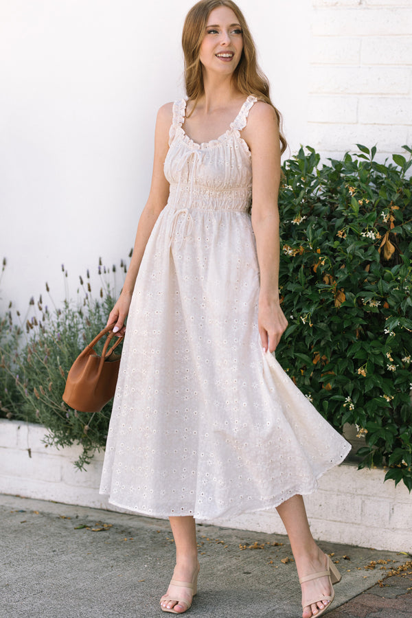 A flowy, pale pink eyelet midi dress with ruffled shoulder straps and a gathered waistline, styled with a tan tote bag and sandals.