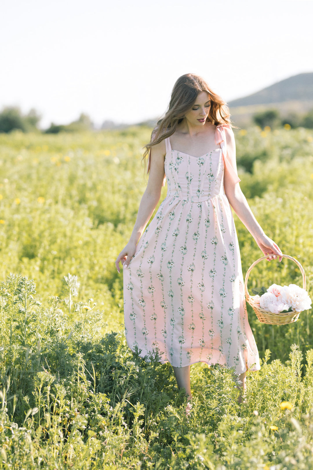 A woman in a light pink floral midi dress holding a basket of white flowers, standing in a field of lush green plants and yellow wildflowers.