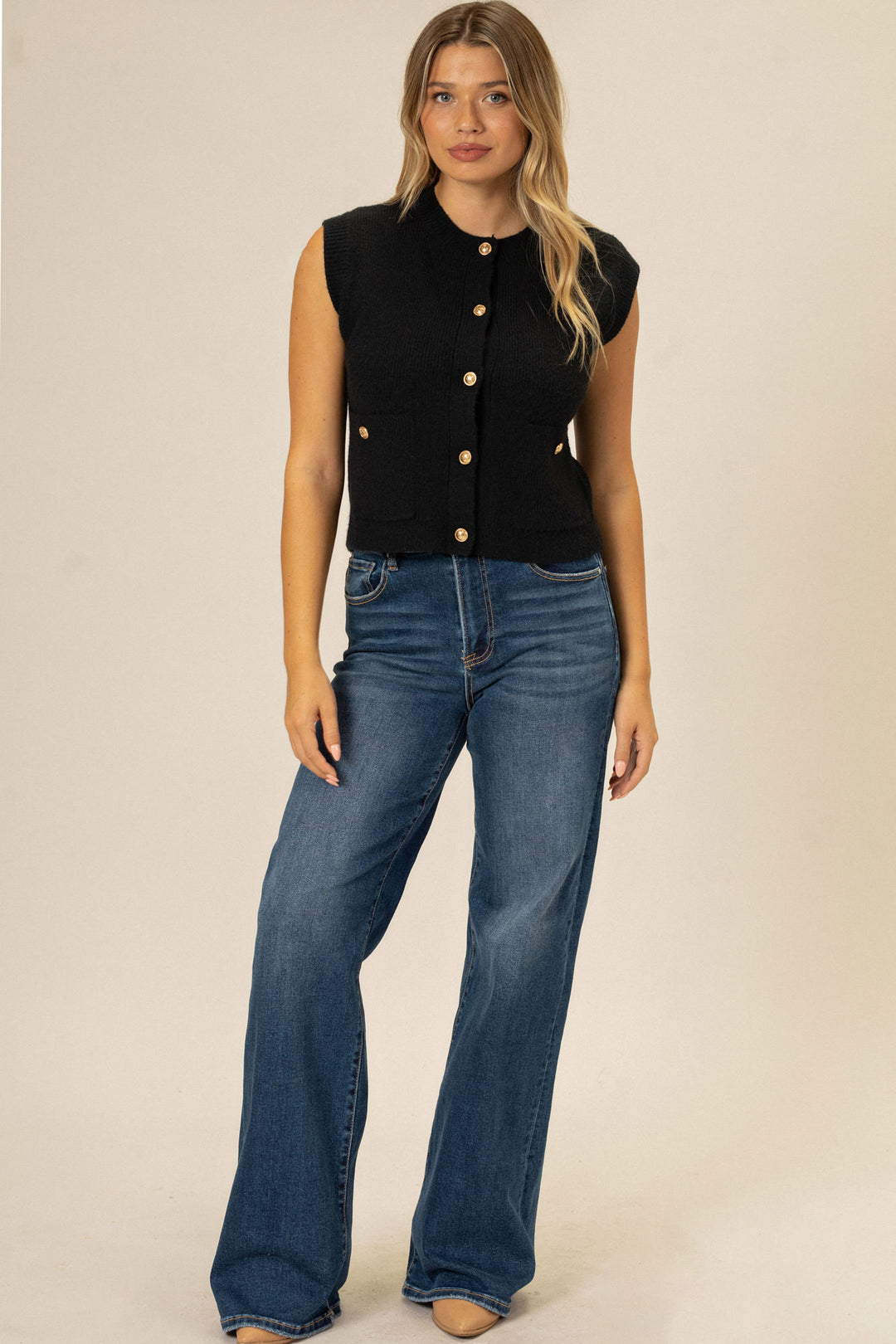 A woman wearing a black knitted sleeveless vest with buttons, paired with high-waisted blue jeans, modeling the Abby Pocket Knit Vest.